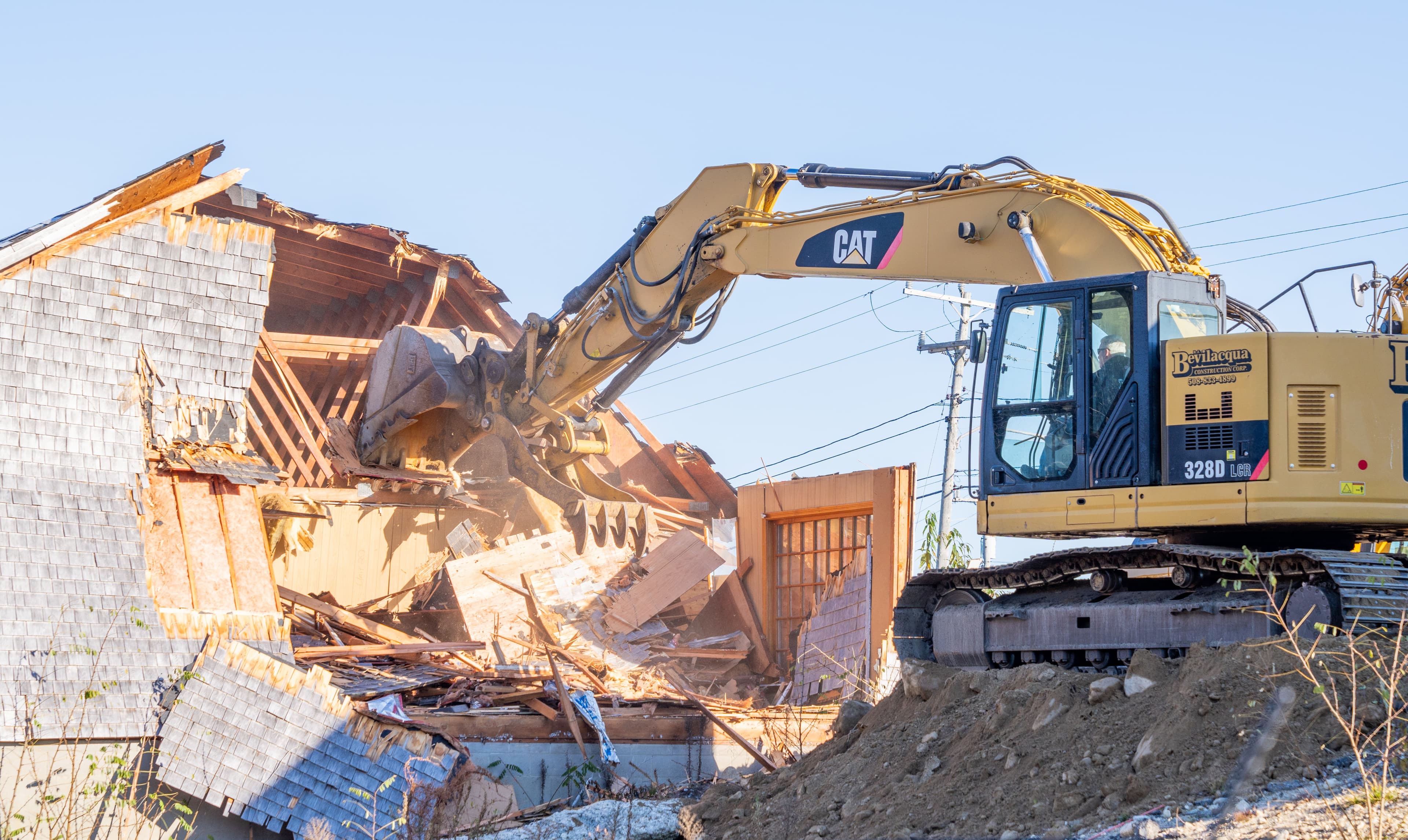 Yellow CAT excavator demolishing a shingle-sided house, its bucket tearing through the wooden structure.