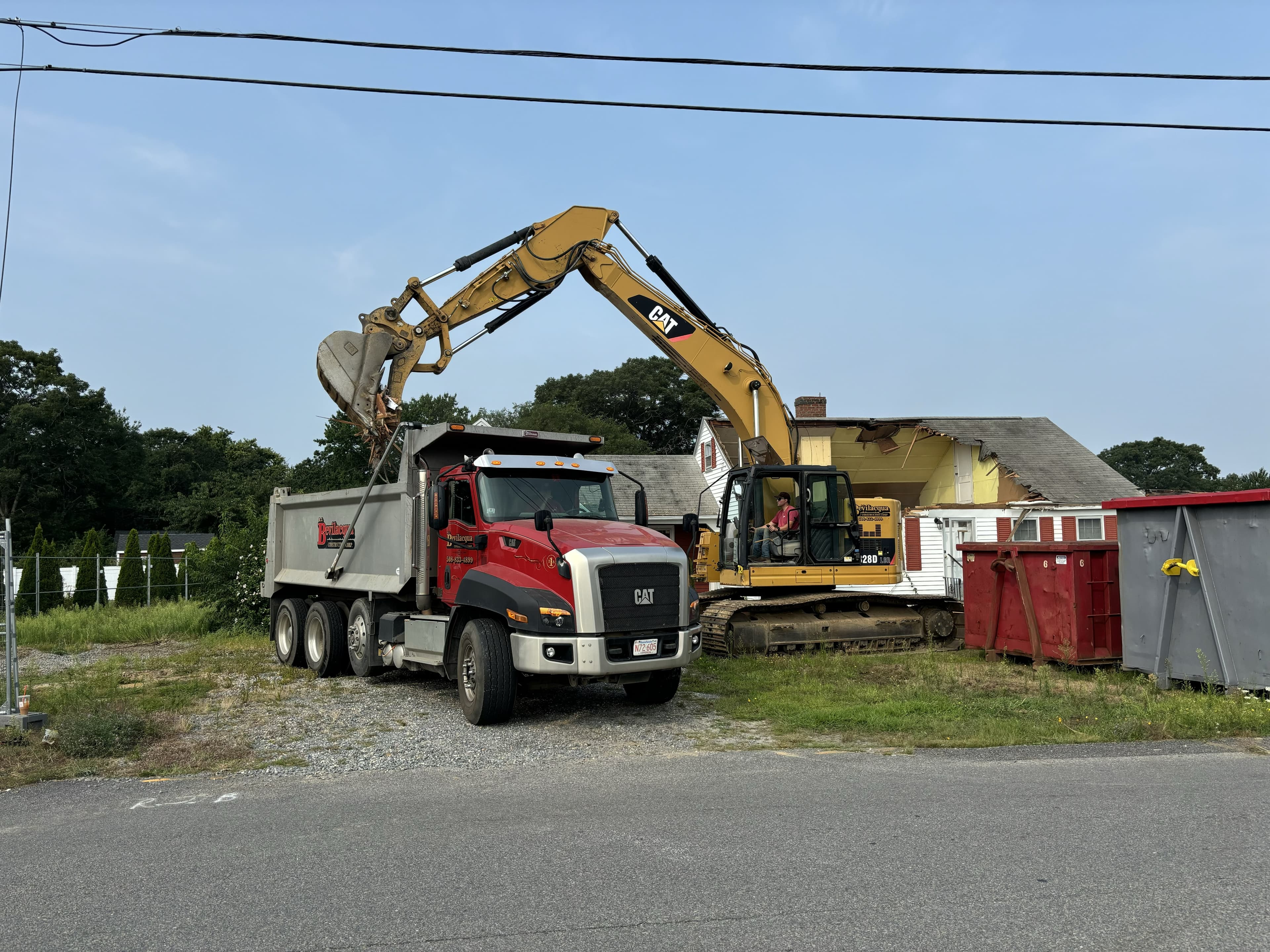 Yellow excavator loading debris into a red dump truck at a residential demolition site.