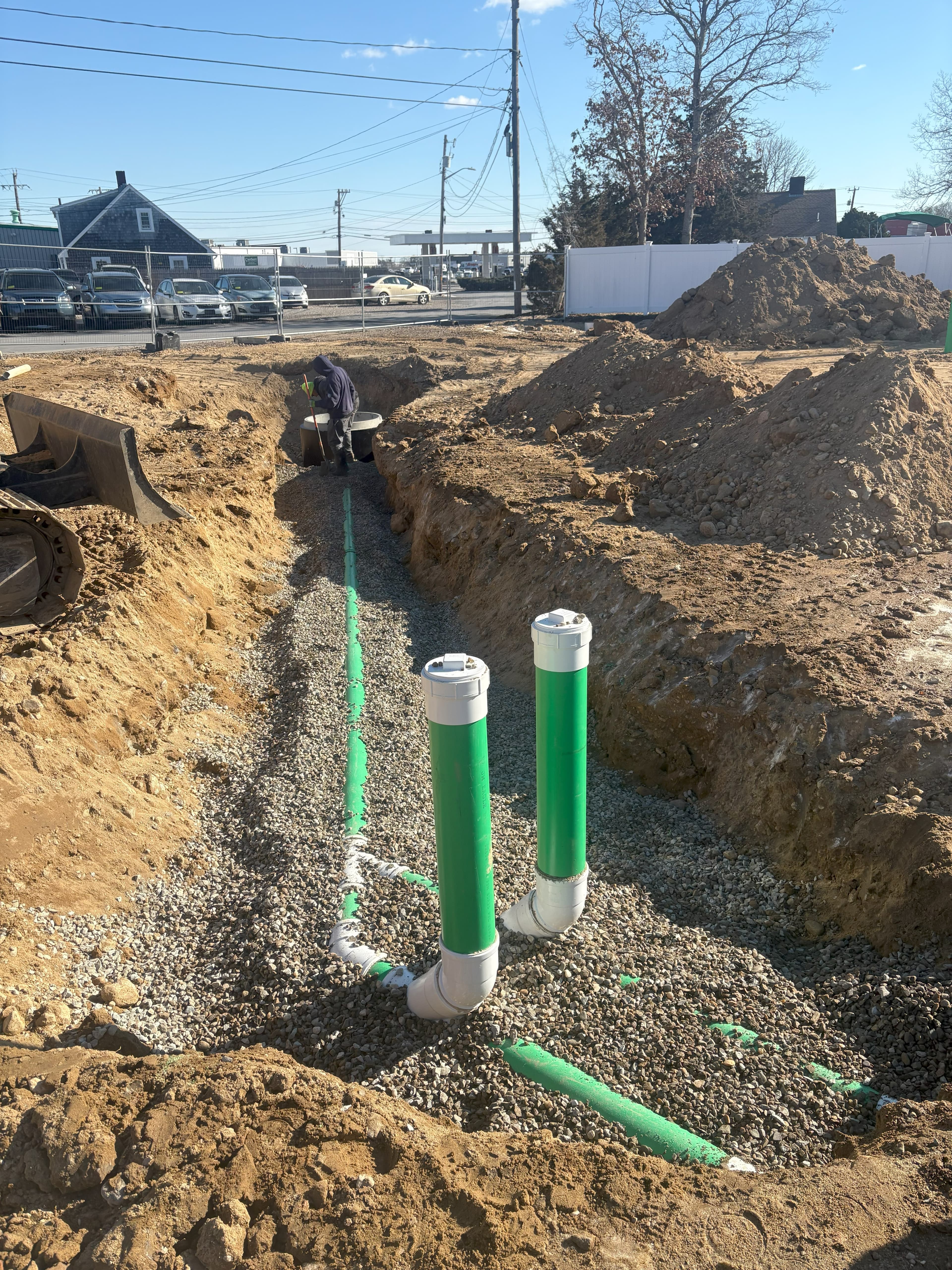 Worker in a deep dirt trench installing green pipes on a bed of gravel.