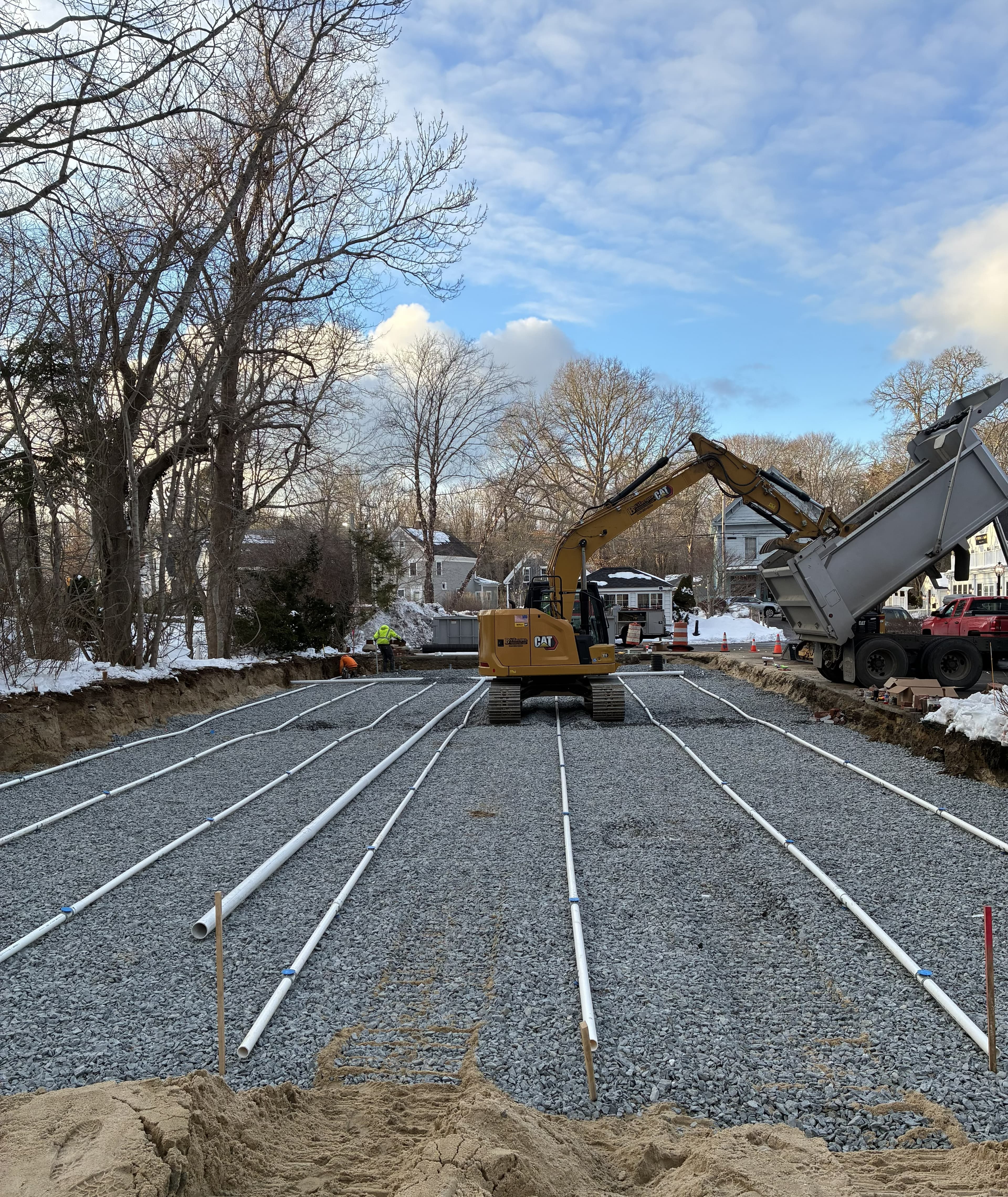 Yellow excavator and dump truck installing white drainage pipes on a gravel construction site.