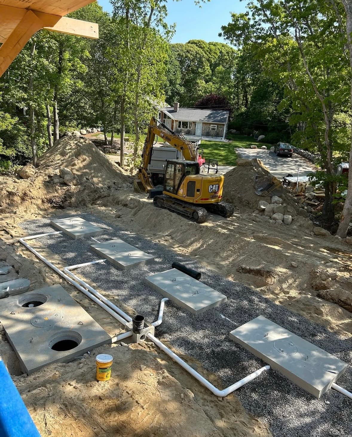 Yellow excavator installing a residential septic system with concrete tanks and white PVC pipes.