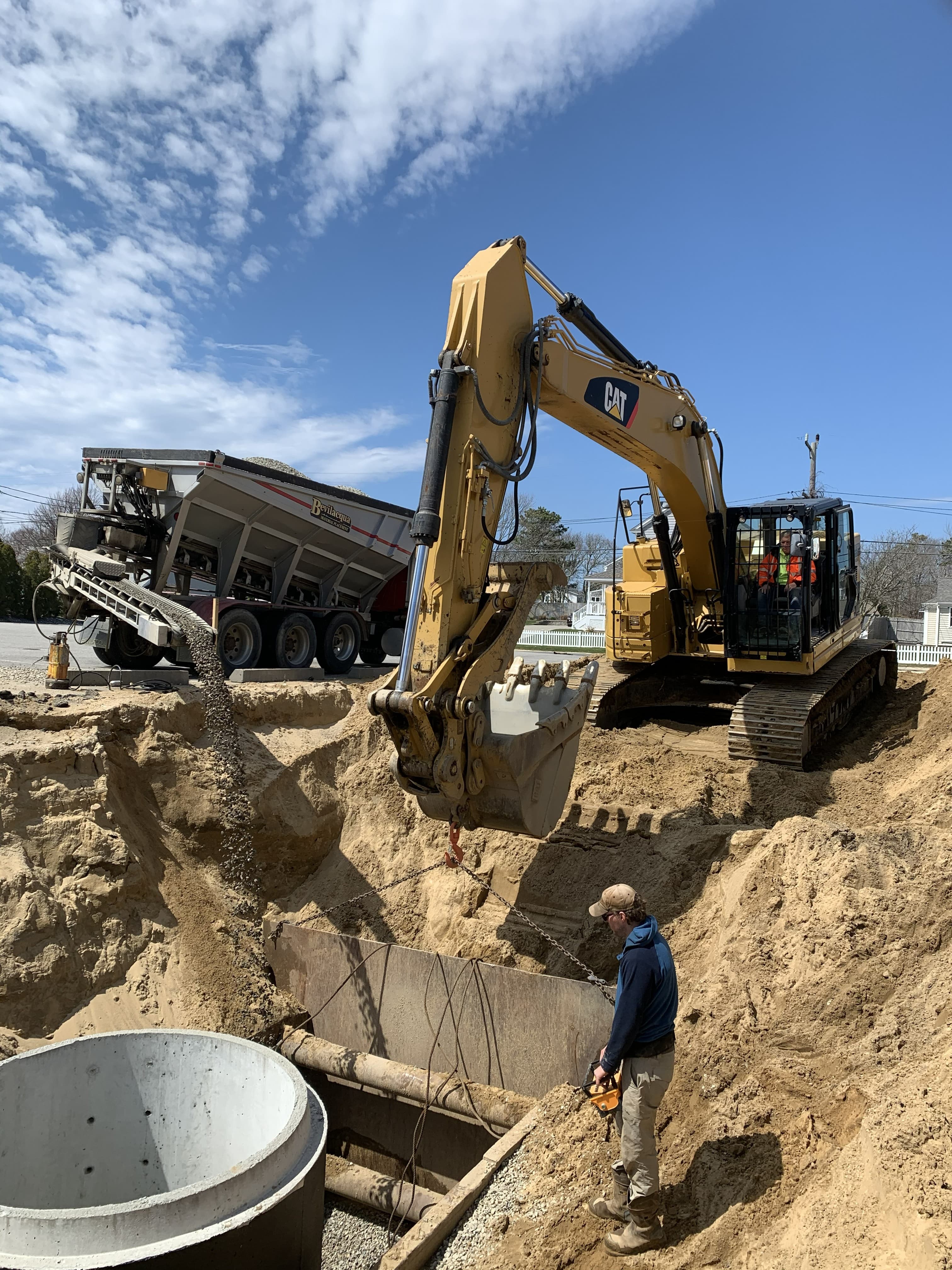 Yellow excavator and truck pouring gravel into a deep trench for underground utility installation.