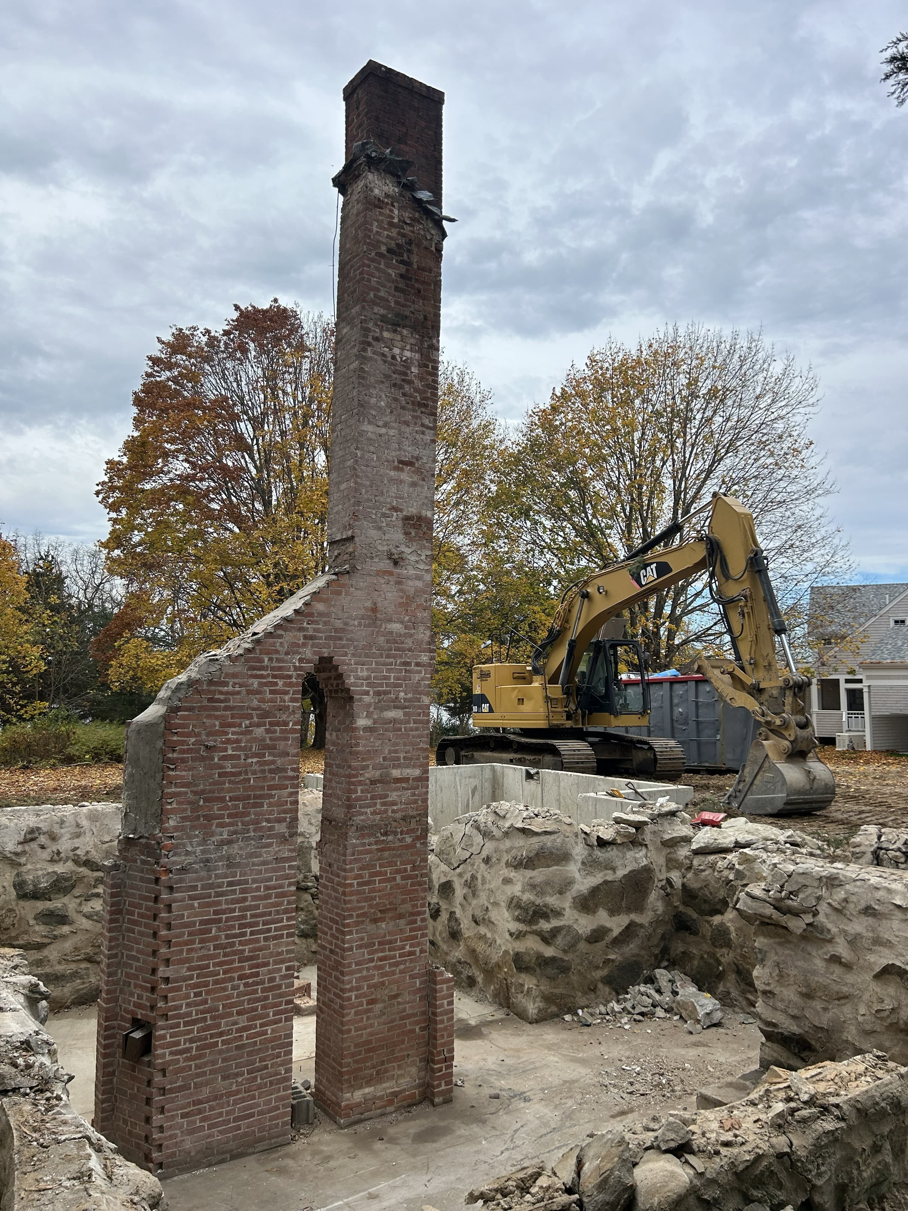 Solitary brick chimney stands amidst stone foundations at a demolition site with a yellow excavator.