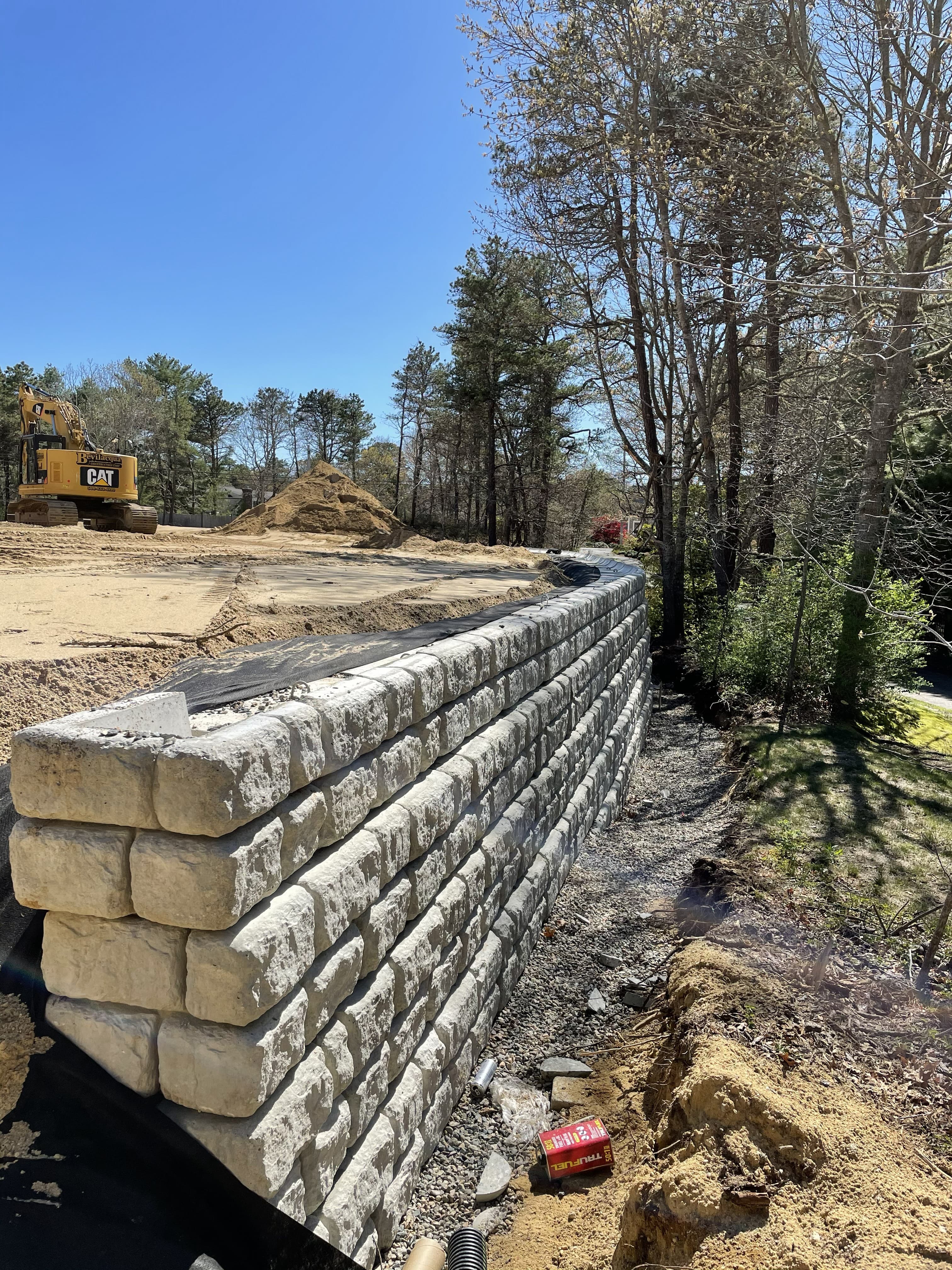 Curved stone block retaining wall under construction with a yellow excavator in the background.